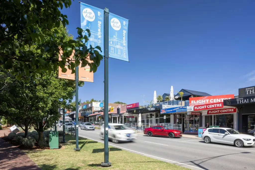 A sunny street scene with cars driving past shops, including a Flight Centre and Thai restaurant. Colorful "Shop Local" banners hang from street poles, and trees line the sidewalk under a clear blue sky.
