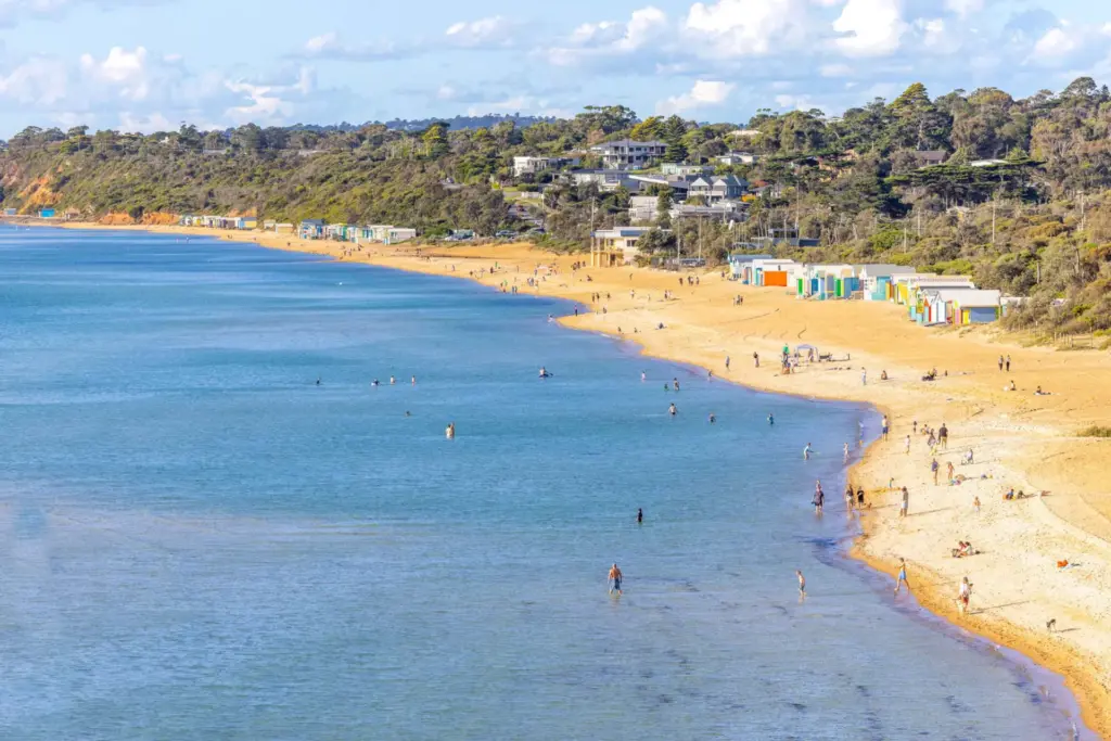 A sandy beach curves along calm blue water with people swimming and walking. Colorful beach huts line the shore, and houses and trees sit on a hill in the background under a partly cloudy sky.
