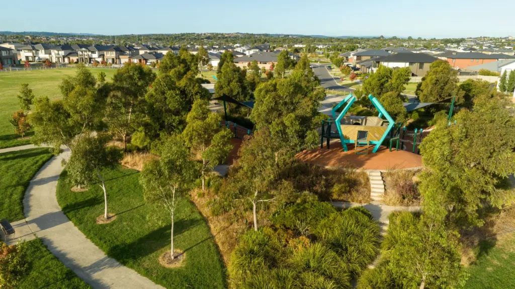 Aerial view of a suburban park with walking paths, green trees, and a modern playground featuring blue geometric climbing structures, surrounded by houses and open grassy areas.